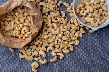 Paper bag and bowl filled with roasted cashew nuts placed on a textured gray surface, with a few nuts scattered around for a natural, appetizing look.