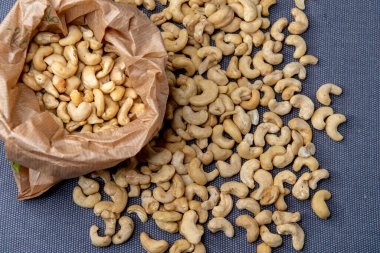 Paper bag filled with roasted cashew nuts placed on a textured gray surface, with a few nuts scattered around for a natural, appetizing look.
