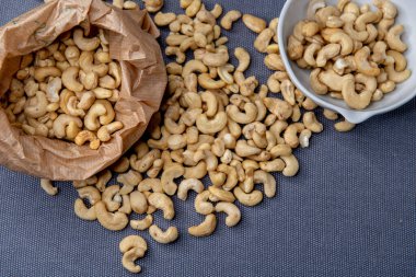 Paper bag and bowl filled with roasted cashew nuts placed on a textured gray surface, with a few nuts scattered around for a natural, appetizing look.