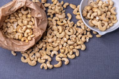 Paper bag and bowl filled with roasted cashew nuts placed on a textured gray surface, with a few nuts scattered around for a natural, appetizing look.