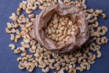 Paper bag filled with roasted cashew nuts placed on a textured gray surface, with a few nuts scattered around for a natural, appetizing look.