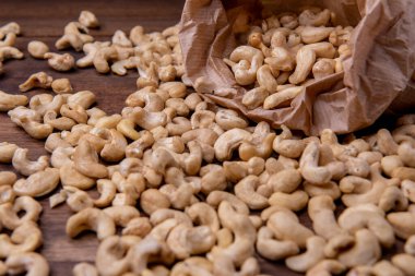roasted cashew nuts placed on a textured brown wooden surface, with a few nuts scattered around for a natural, appetizing look. paper bag filled with roasted cashew nuts