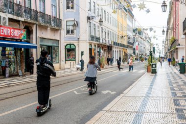 İki kişi Lizbon caddesinde elektrikli scooter kullanıyor. Renkli binalar, dükkanlar ve dekorasyonlarla dolu. Şehir merkezinin canlı atmosferini yakalıyorlar..