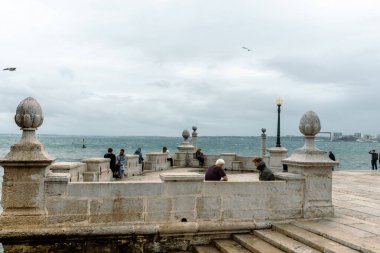Lizbon. Dağılmış ziyaretçilerin bulutlu bir gökyüzünün altındaki dalgalı denize bakıp sakin ama rüzgârlı bir kıyı atmosferini yakaladığı taştan bir rıhtım geçidi..