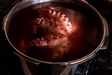 Sobanın tepesindeki paslanmaz çelik bir tencerede kaynayan ahtapotun yakın çekiminde detaylı doku, vakum bardakları ve mutfak ortamında deniz ürünleri pişirme süreci görülüyor..