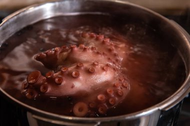 Sobanın tepesindeki paslanmaz çelik bir tencerede kaynayan ahtapotun yakın çekiminde detaylı doku, vakum bardakları ve mutfak ortamında deniz ürünleri pişirme süreci görülüyor..