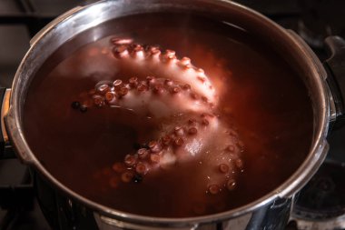 Sobanın tepesindeki paslanmaz çelik bir tencerede kaynayan ahtapotun yakın çekiminde detaylı doku, vakum bardakları ve mutfak ortamında deniz ürünleri pişirme süreci görülüyor..