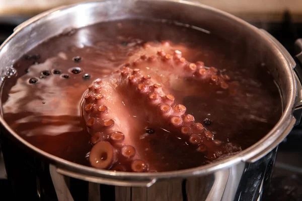 Sobanın tepesindeki paslanmaz çelik bir tencerede kaynayan ahtapotun yakın çekiminde detaylı doku, vakum bardakları ve mutfak ortamında deniz ürünleri pişirme süreci görülüyor..