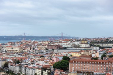 Lizbon 'un tarihi Alfama bölgesinin panoramik manzarasında tepenin üstünde Sao Jorge Şatosu, kırmızı kiremit çatıları olan renkli binalar ve arka planda Tagus Nehri üzerindeki 25 de Abril asma köprüsü yer alıyor..