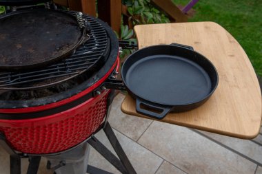 Ahşap rafa dökme demir bir tavayla seramik bir kömür ızgarasının yakın çekimi. Bahçede barbekü aksesuarları ve doğal ışık olan açık hava yemekleri..