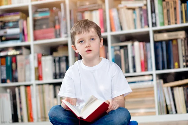 Menino com livro vermelho na biblioteca — Fotografia de Stock