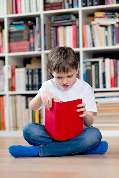 Weinig jongenskind lezen van een rode boek in de bibliotheek. — Stockfoto