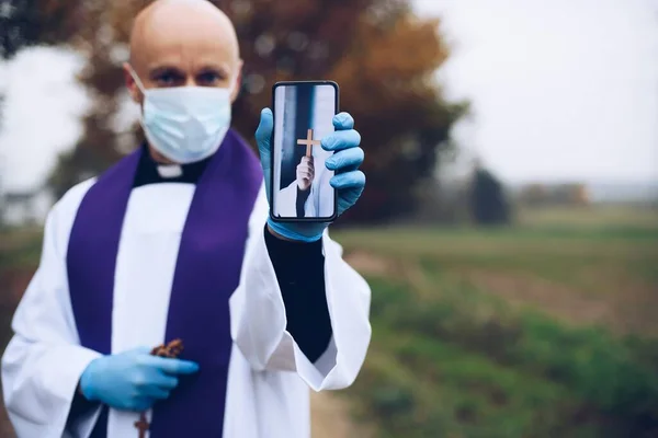 Sacerdote em uma máscara médica e luvas de proteção mantém um telefone celular com cruz. — Fotografia de Stock