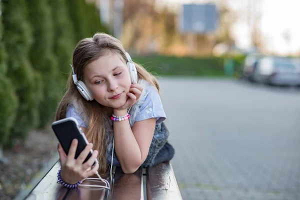 La chica está tumbada en el banco y escuchando música desde el teléfono —  Fotos de Stock