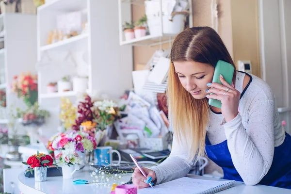 Young woman florist noting client order during mobile phone conversation in a flower shop — Stock Photo, Image
