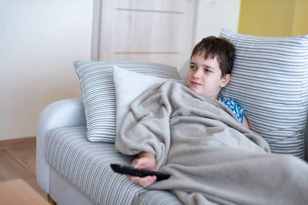 A young boy is lying on the sofa under a blanket, with the remote control in his hand, watching TV. — Stock Photo, Image