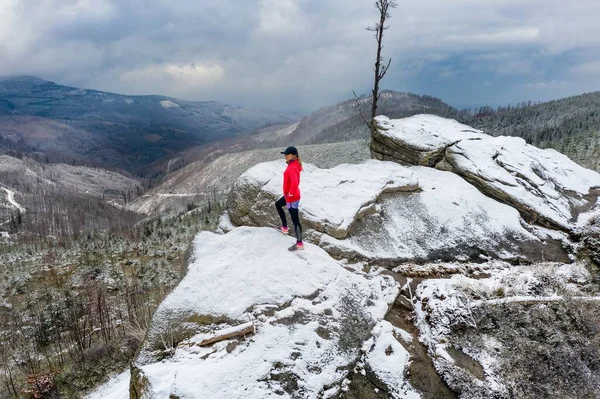 Εναέρια άποψη για ένα κορίτσι δρομέας σε ένα κόκκινο φούτερ στέκεται σε ένα χιονισμένο βράχο θαυμάζοντας την ομορφιά των βουνών σε μια συννεφιασμένη μέρα. — Φωτογραφία Αρχείου