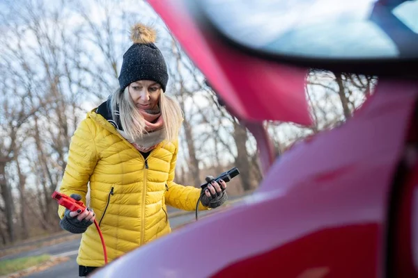 Beautiful blonde woman in yellow jacket trying to start her broken car with jumper cables. — Stockfoto