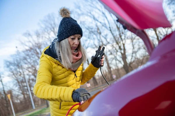 Beautiful blonde woman in yellow jacket trying to start her broken car with jumper cables. — Stockfoto