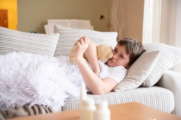 Droevige zieke jongen ligt op de bank knuffelend zijn teddybeer. — Stockfoto