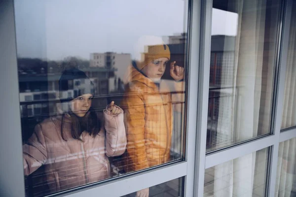 Dos niños están de pie junto a la ventana y tristemente mirando afuera durante la cuarentena o el aislamiento debido a la pandemia. — Foto de Stock