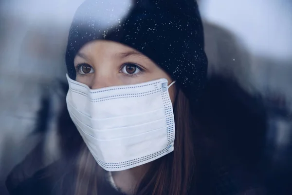 A pretty girl is standing by the window in a protective mask.