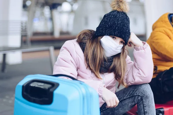 Ein trauriges Mädchen mit Schutzmaske sitzt auf dem Bahnsteig und träumt vom Urlaub. — Stockfoto