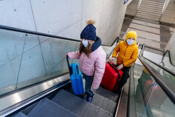 スーツケース付きの保護マスクを着た子供たちがエスカレーターで駅のホームに上がります。パンデミックの時. — ストック写真