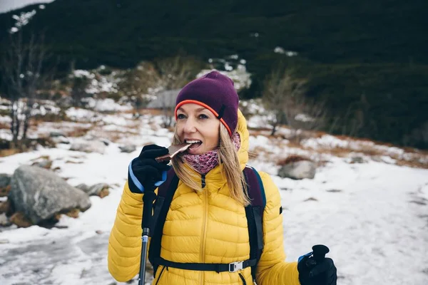 A blonde tourist is eating chocolate on a hiking trail in the mountains.