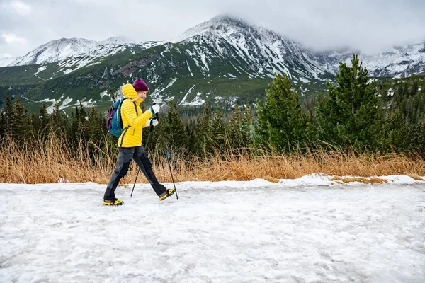 一个金发碧眼的游客穿着一件黄色的外套走在高山上的冬季小径上. — 图库照片