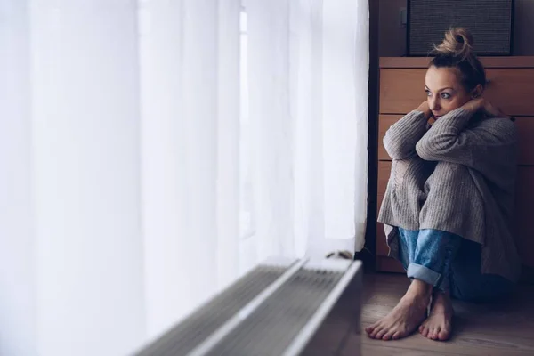 Mladá smutná žena sedí na podlaze v obývacím pokoji a dívá se z okna.. — Stock fotografie