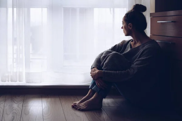 Mujer triste sentada en el suelo en su sala de estar mirando por la ventana. — Foto de Stock