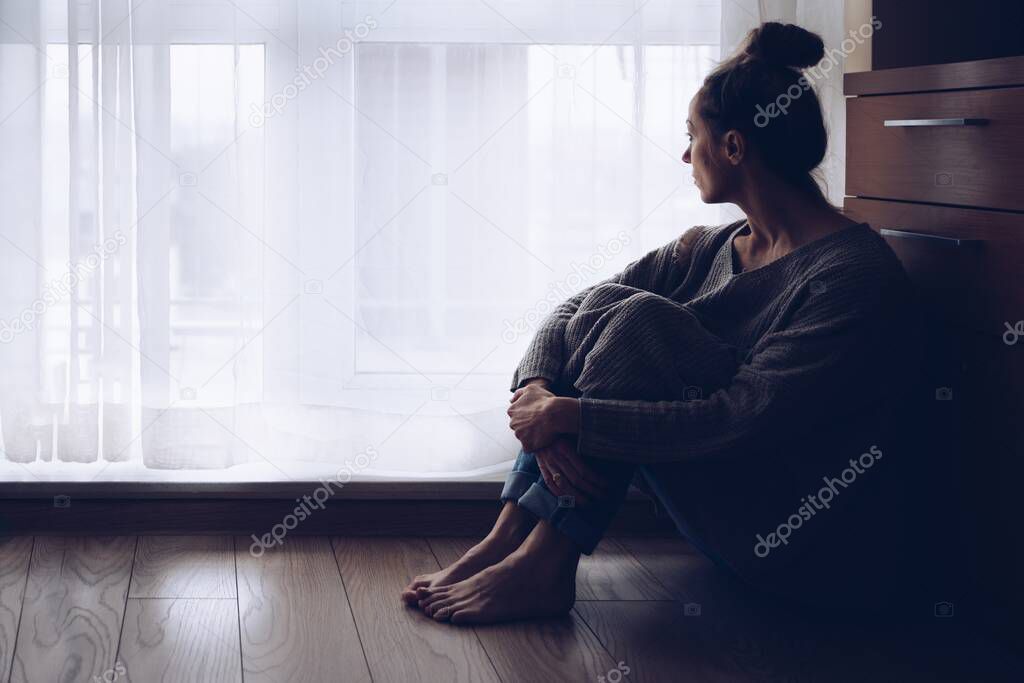 Sad woman sitting on the floor in her living room looking out the window.