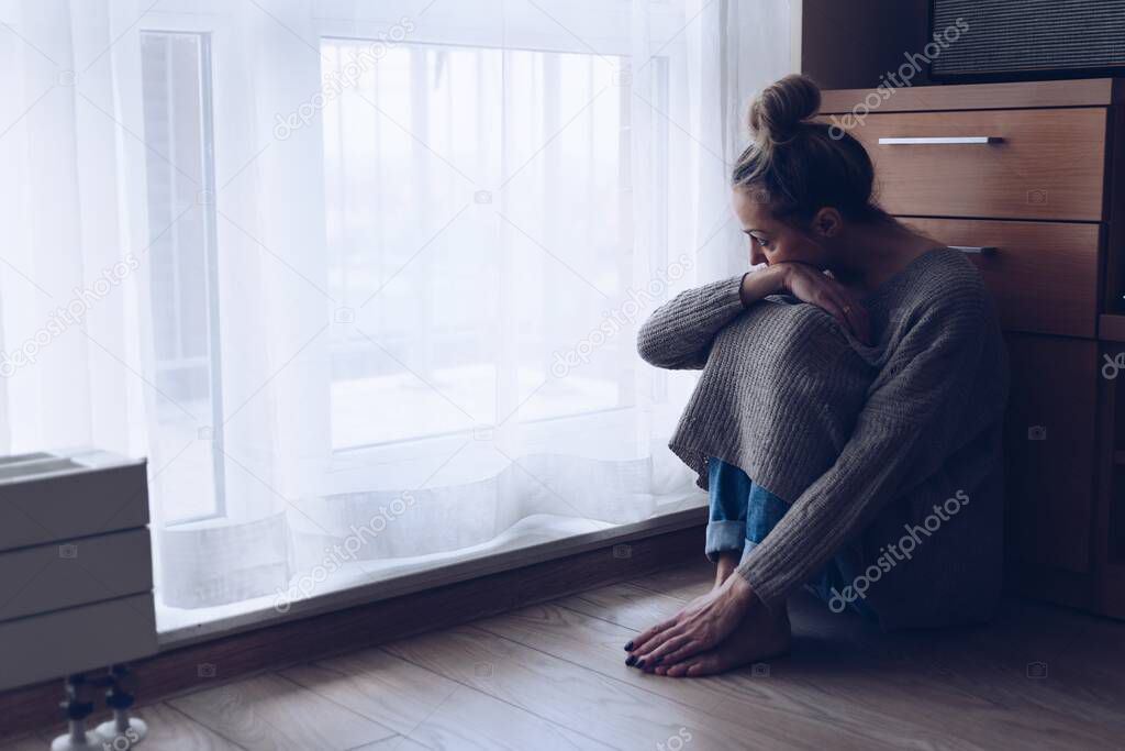 A devastated sad woman is sitting on the floor in her living room looking at the window.