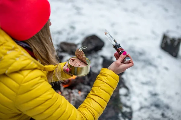 En kvinna i gul jacka med konserverad mat vid en brasa på en fjällstig. — Stockfoto