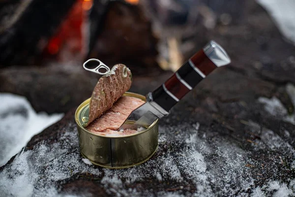 Open can of meat. Knife stuck inside. A burning fire.