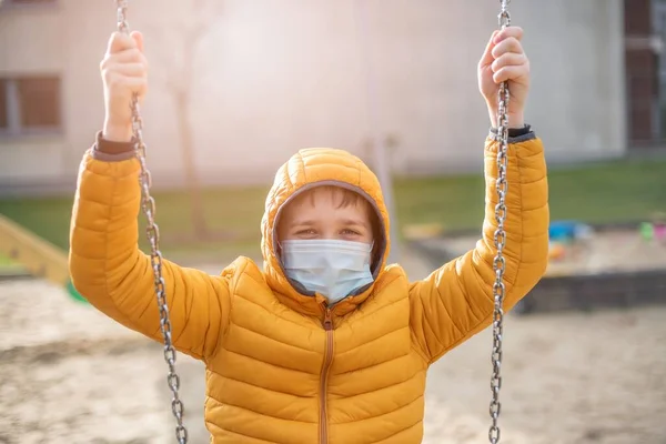 Chłopiec w żółtej kurtce w masce ochronnej podczas pandemii na placu zabaw. — Zdjęcie stockowe