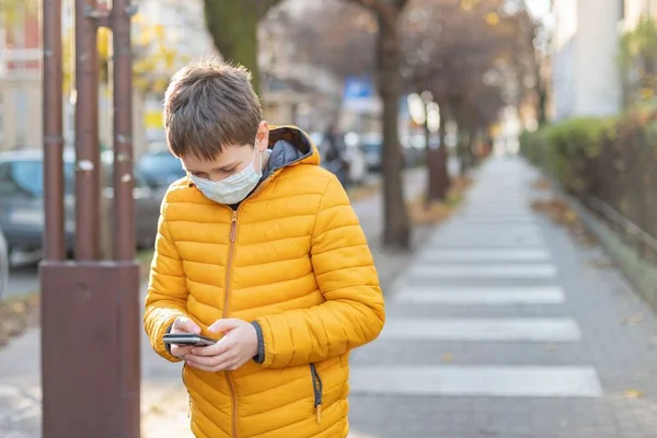 Un niño con una máscara protectora está en la calle sosteniendo un teléfono en su mano. — Foto de Stock