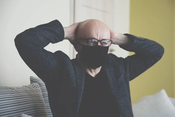 Um homem está sentado em um sofá usando uma máscara protetora agarra a cabeça. — Fotografia de Stock