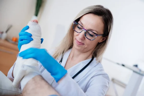 Enfermeira profissional sorridente com óculos é enfaixar a perna. Tratamento de ossos partidos. — Fotografia de Stock