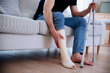 Kırık bacaklı genç bir adam, bir kazadan sonra bacağında beyaz alçıyla kanepede oturuyor..