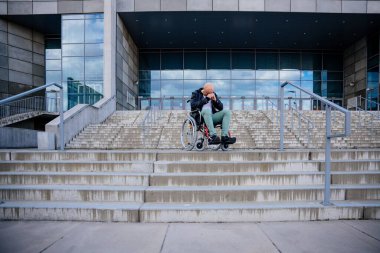 Üzgün, sakat bir adam alnına dokunuyor ve merdivenlerin önünde tekerlekli sandalyede otururken üzgün görünüyor..