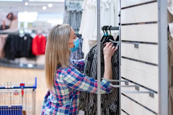 Mujer con mascarillas protectoras en una tienda de ropa eligiendo ropa nueva en el departamento de venta. —  Fotos de Stock