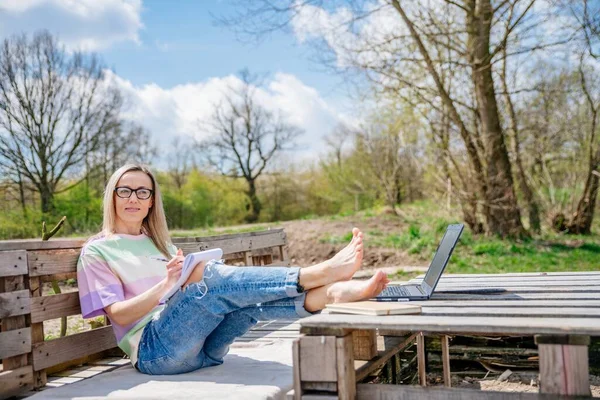 Piękna blondynka pracująca na zewnątrz. Trzyma jego stopy na stole paletowym obok laptopa.. — Zdjęcie stockowe