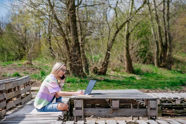 Blondynka pracuje na zewnątrz za pomocą laptopa i telefonu komórkowego. — Zdjęcie stockowe