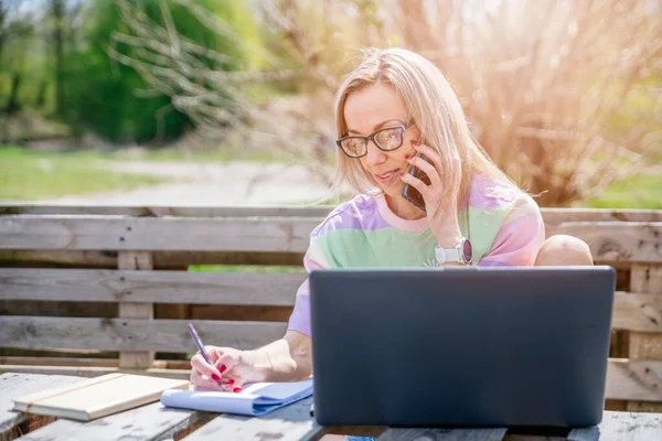 Die umtriebige Blondine arbeitet bei schönem Sonnenschein aus der Ferne. Sie telefoniert. Arbeiten im Freien. — Stockfoto