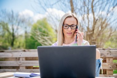 Gözlüklü kadın güneşli bir günde dışarıda dizüstü bilgisayar ve telefon kullanıyor..