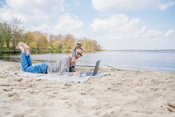 Mulher trabalha ao ar livre no laptop. Ela está deitada na praia em um cobertor e falando ao telefone com klient. — Fotografia de Stock