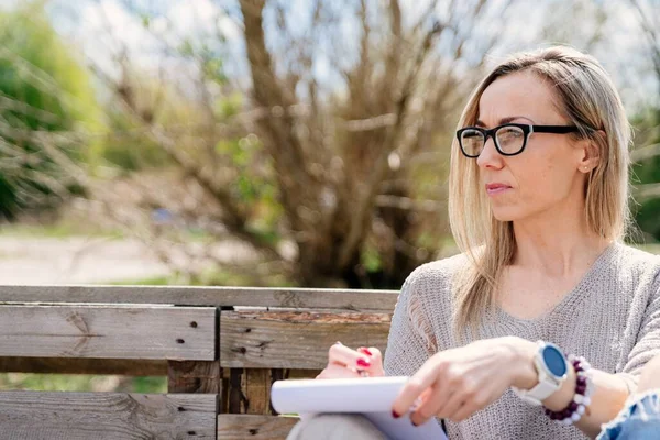 Encantadora mujer hipster en gafas escribiendo reseña interesante sobre viajes en notebook al aire libre. —  Fotos de Stock