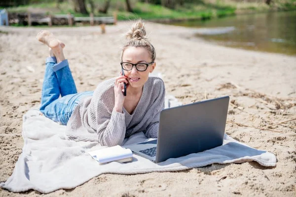 Lächelndes blondes Mädchen arbeitet draußen am Laptop. Auf einer Decke am Strand liegend, telefonierend. — Stockfoto